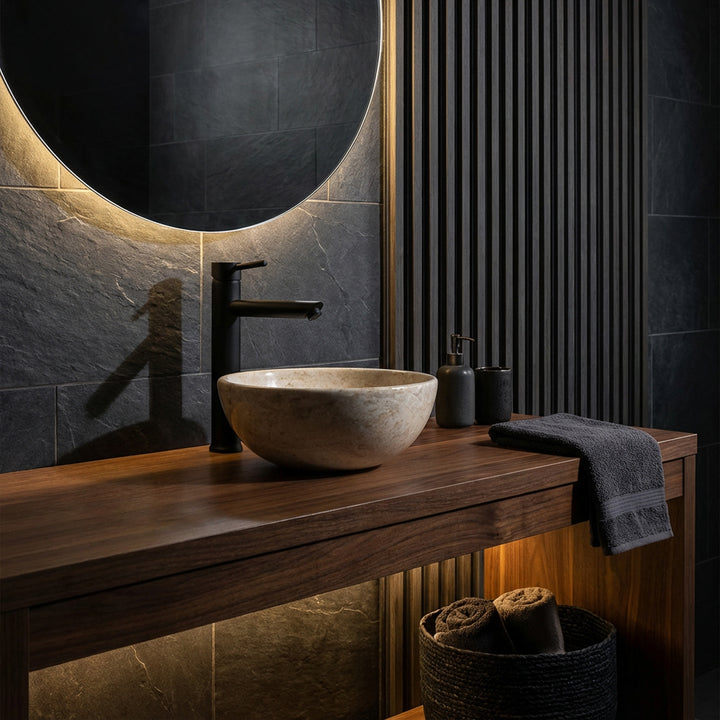 Rundes Aufsatzwaschbecken aus hellem Naturstein auf dunklem Holzwaschtisch mit schwarzer Armatur im eleganten Badambiente