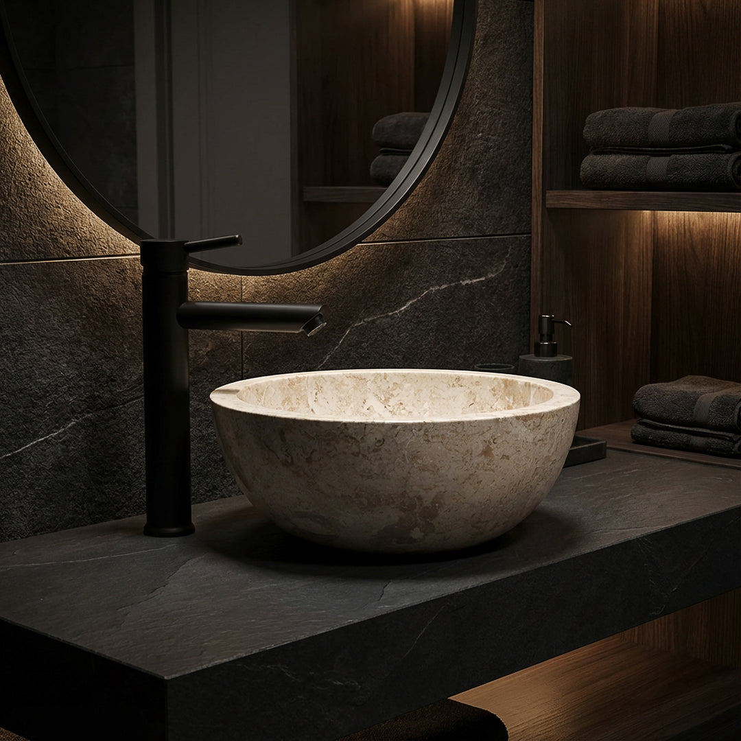 Rundes Aufsatzbecken aus hellem Marmor auf dunkler Steinplatte mit schwarzer Armatur im eleganten Badambiente