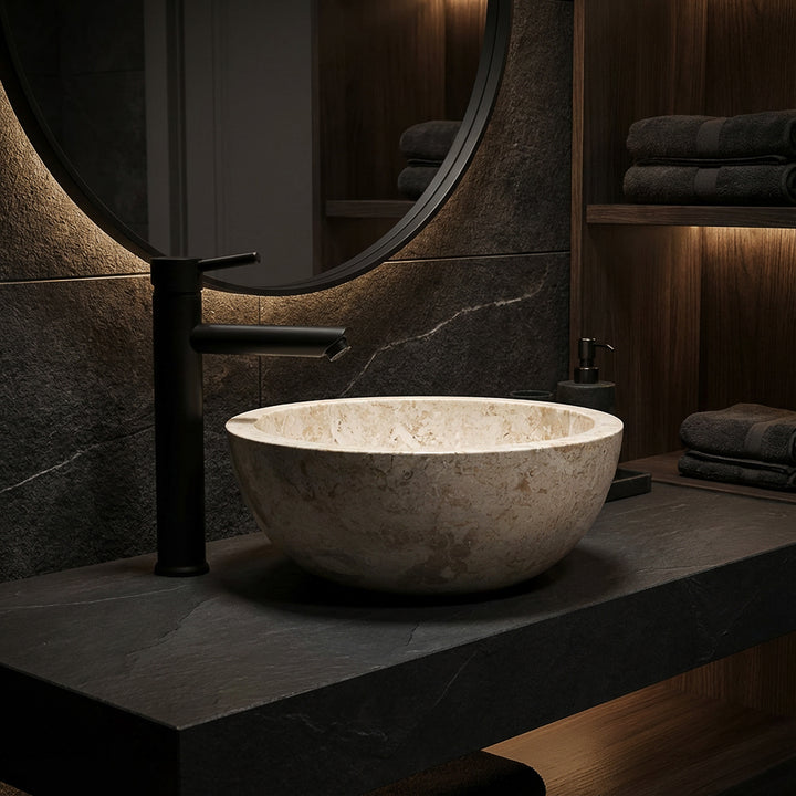 Rundes Aufsatzbecken aus hellem Marmor auf dunkler Steinplatte mit schwarzer Armatur im eleganten Badambiente