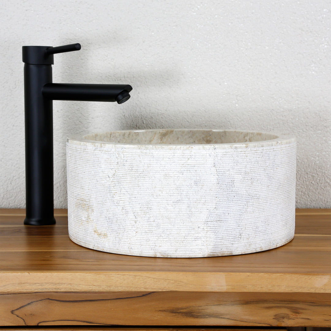 Rundes Aufsatzwaschbecken aus hellem Naturstein mit feiner Linienstruktur, kombiniert mit schwarzer Standarmatur.