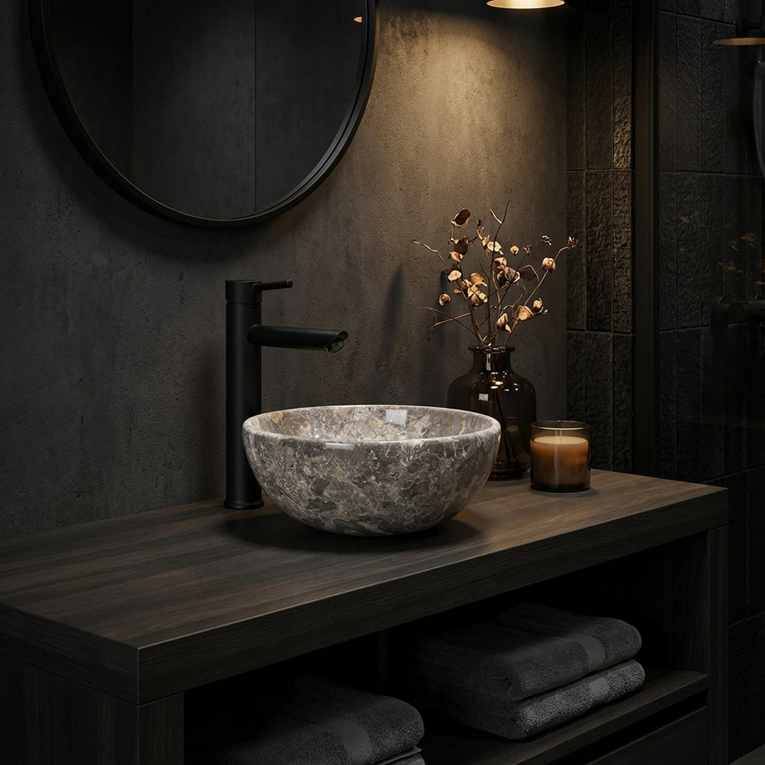 Rundes Naturstein Aufsatzwaschbecken mit graubrauner Maserung auf dunkler Holzkonsole im stimmungsvoll beleuchteten Badezimmer, kombiniert mit schwarzer Armatur, Spiegel und dezenten Accessoires