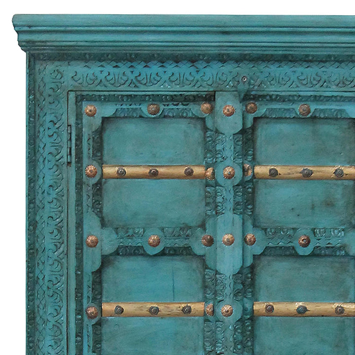 Detail der türkisfarbenen Schrankfront mit geschnitzten Ornamenten, goldfarbenen Leisten und Beschlägen.