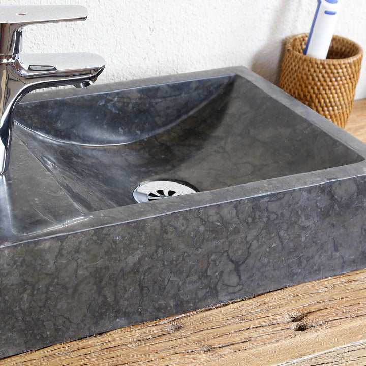 Eckiges Naturstein Aufsatzbecken mit dunkler Maserung in Detailansicht, mit Edelstahl-Abfluss und verchromter Armatur auf Holzplatte