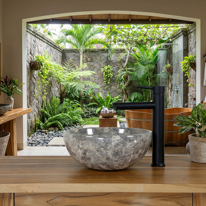 Rundes Naturstein Waschbecken mit graubrauner Maserung auf Holzwaschtisch im offenen Spa-Badezimmer mit Blick in einen grünen Innenhof, kombiniert mit schwarzer Standarmatur, Natursteinmauer und Holzbadewanne