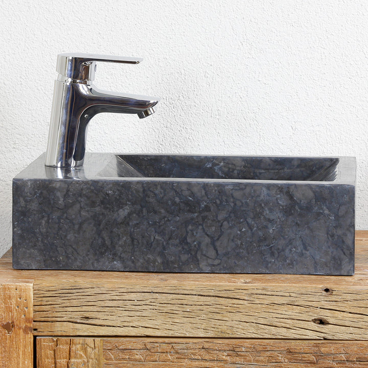Eckiges Aufsatzbecken aus Marmor mit dunkler Maserung auf Holzkonsole, kombiniert mit verchromter Armatur vor heller Wand
